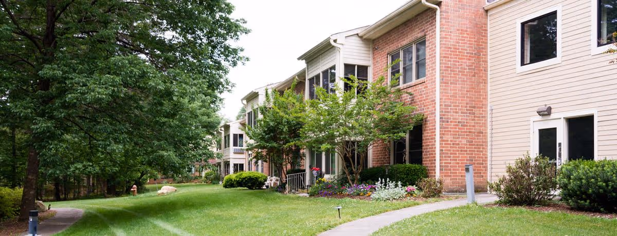 Exterior view of a senior living facility building with brick and siding walls, surrounded by green grass, trees, and landscaped bushes and flowers. A paved walkway curves through the lawn in front of the building.