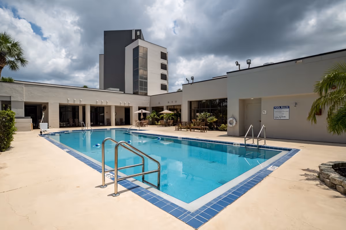 Outdoor swimming pool area at a senior living facility with clear blue water, surrounded by a concrete deck. There are metal handrails for pool entry, lounge chairs, tables with umbrellas, and some greenery including palm trees. The building surrounding the pool is white with large windows and a tall multi-story section in the background under a cloudy sky.