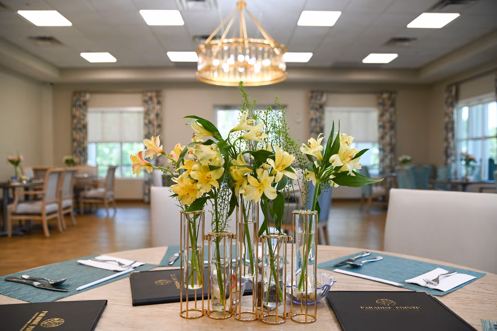 A dining room in an assisted living facility with a round table in the foreground decorated with a centerpiece of yellow flowers in glass vases with gold holders. The table is set with placemats, silverware, and menus. In the background, there are more tables and chairs, windows with patterned curtains, and a chandelier hanging from the ceiling.