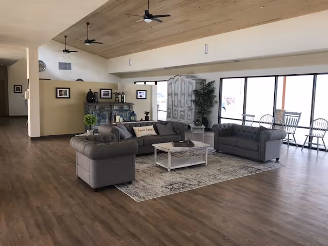 A spacious living room area with wooden flooring and a light wooden ceiling. The room features three gray tufted sofas arranged around a white coffee table on a patterned rug. In the background, there is a decorative cabinet, a large plant, and several framed pictures on the walls. Large windows let in natural light, and there are ceiling fans above.