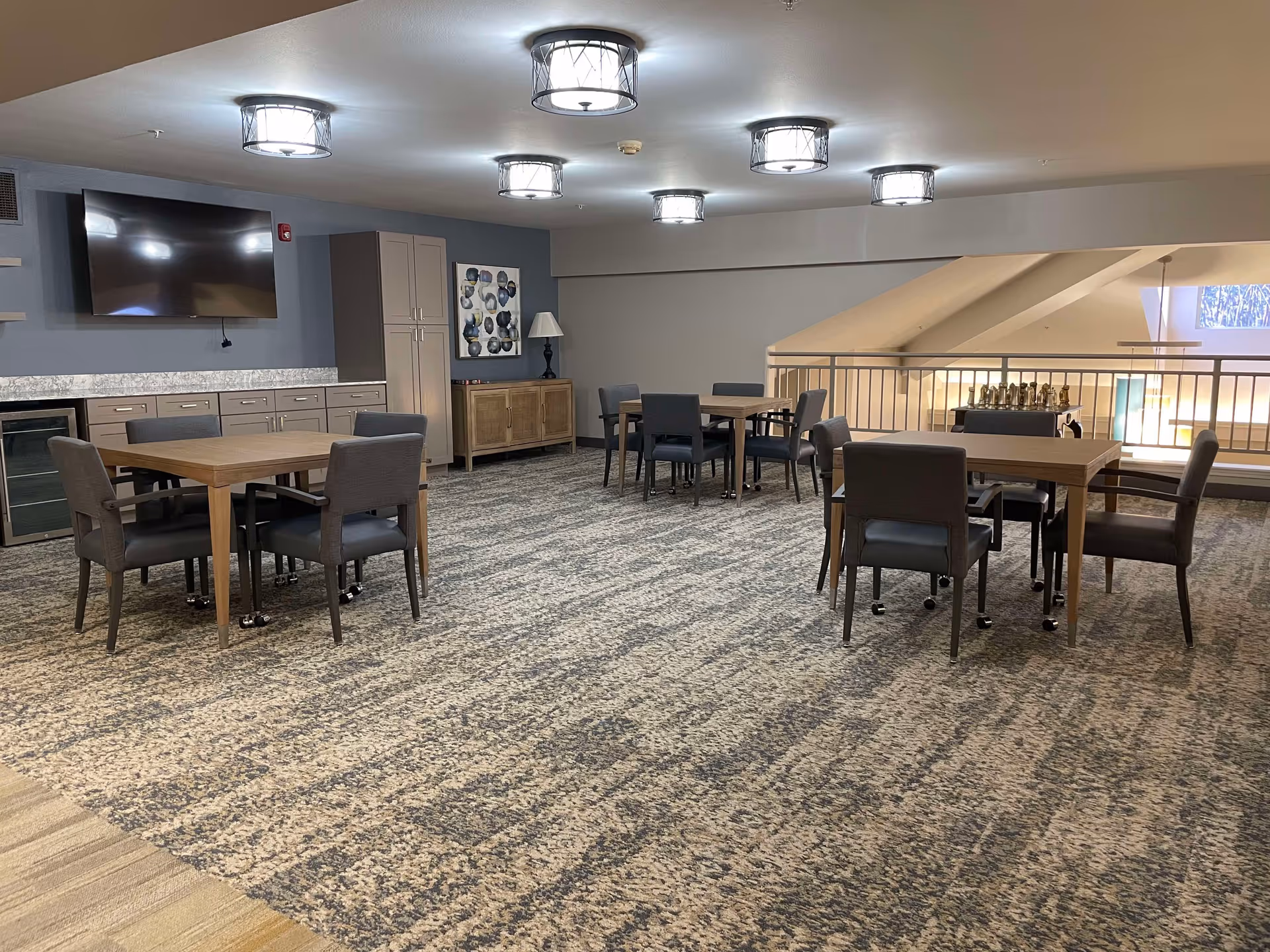 A spacious common area with multiple wooden tables and gray upholstered chairs on a patterned carpet. The room features modern ceiling lights, a large flat-screen TV mounted on a blue-gray wall, cabinets, a lamp on a sideboard, and a railing overlooking a lower level with natural light coming through windows.