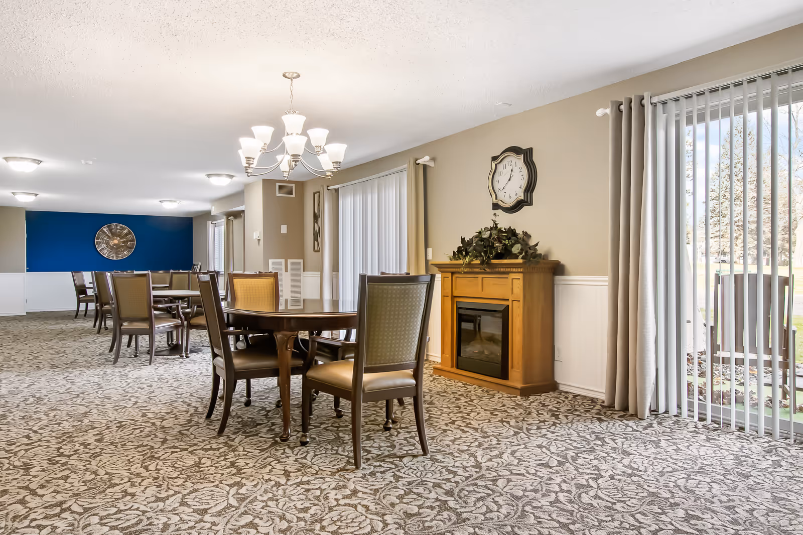 Spacious communal dining room with round tables and chairs, a chandelier, a decorative fireplace cabinet, and sliding glass doors to the outside.