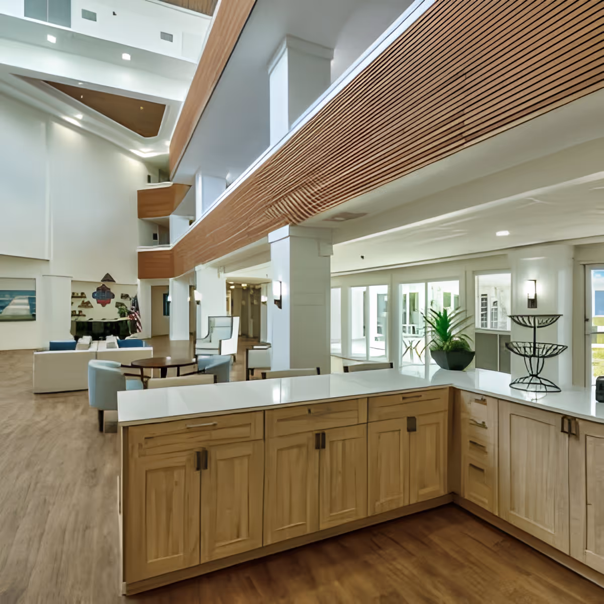 Spacious and modern senior living facility common area with light wood cabinetry in the foreground, comfortable seating with chairs and sofas, a piano, and large windows allowing natural light to fill the space.