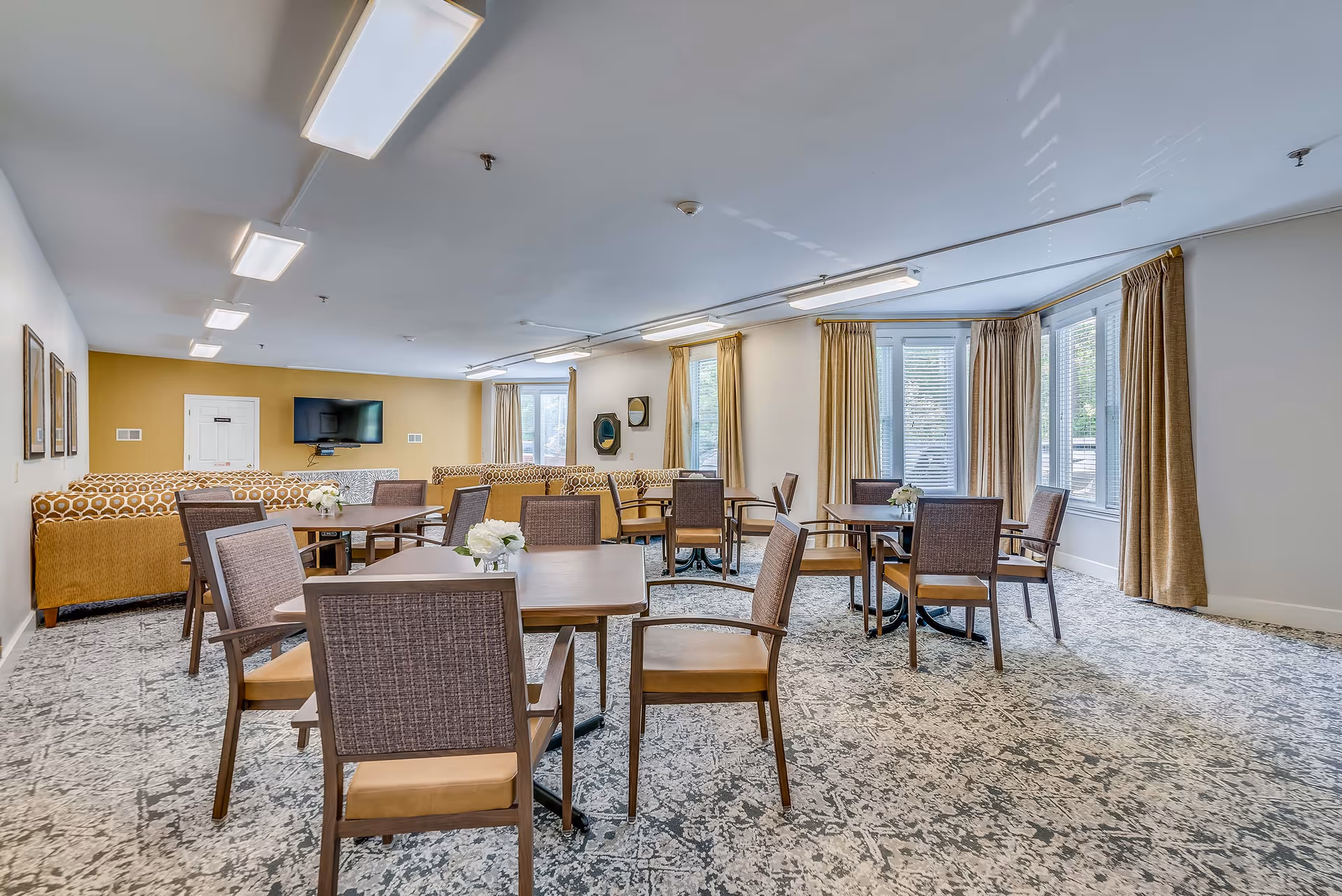 A spacious common area in a retirement residence featuring several square wooden tables each surrounded by four chairs with cushioned seats and woven backs. The room has large windows with beige curtains allowing natural light to fill the space. In the background, there are patterned yellow sofas arranged facing a wall-mounted flat-screen TV on a mustard yellow accent wall. The floor is covered with a patterned carpet in shades of gray and white, and the ceiling has multiple rectangular fluorescent light fixtures.