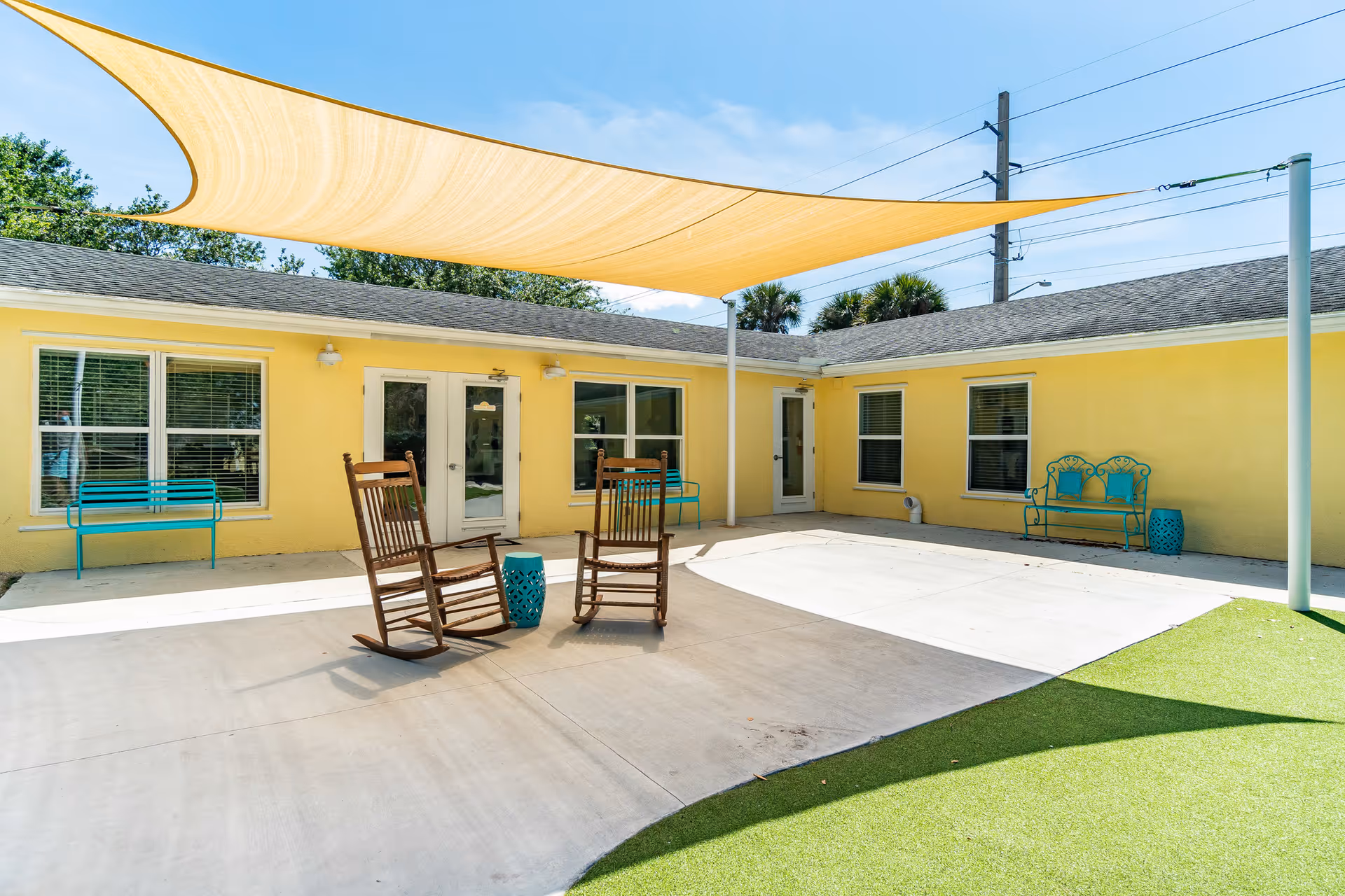 Outdoor courtyard area of Pineapple Garden Assisted Living with two wooden rocking chairs and a small blue side table on a concrete patio. The building walls are painted yellow with several windows and doors. There are turquoise benches along the walls and a large beige shade sail overhead providing partial shade. Green artificial turf is visible on the right side of the image under a clear blue sky.