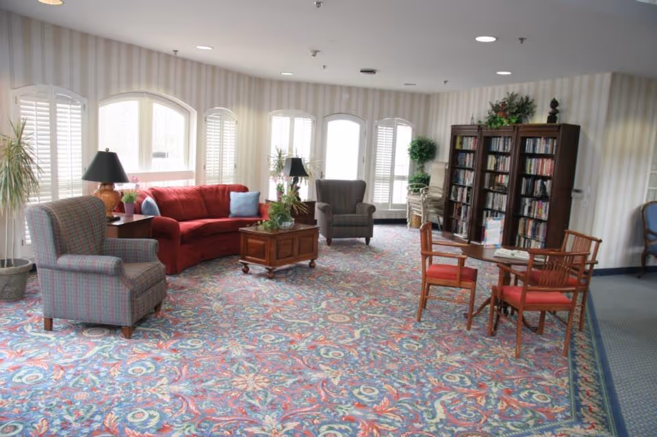 Bright communal living room with patterned carpet, a red sofa, armchairs, coffee table and bookshelves near arched windows.
