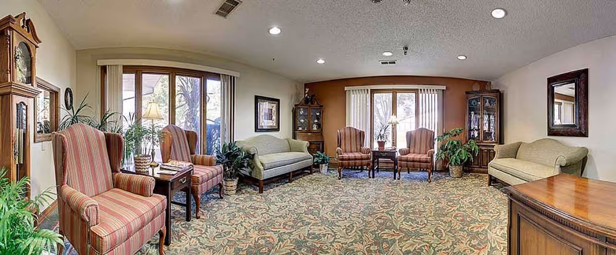 A spacious living room with patterned carpet, two green sofas, four striped armchairs, wooden side tables, potted plants, a grandfather clock, and large windows with vertical blinds letting in natural light.
