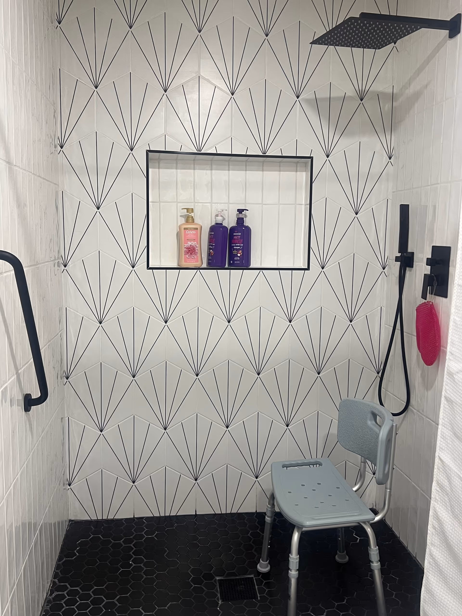 Accessible tiled shower with patterned white tiles, built-in niche holding three bottles, black fixtures, grab bar, and a gray shower chair.