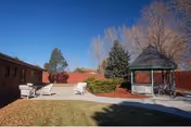 Sunlit outdoor patio area with white chairs, a gazebo, and a red-brick building.