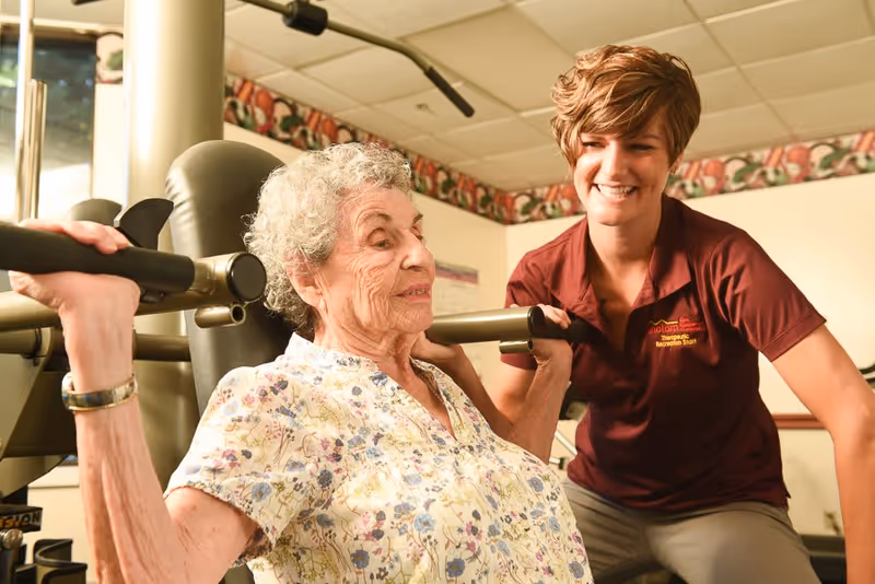 An elderly woman with curly gray hair wearing a floral blouse is using a weight machine for exercise, assisted by a smiling young female therapist in a maroon shirt in a well-lit room.