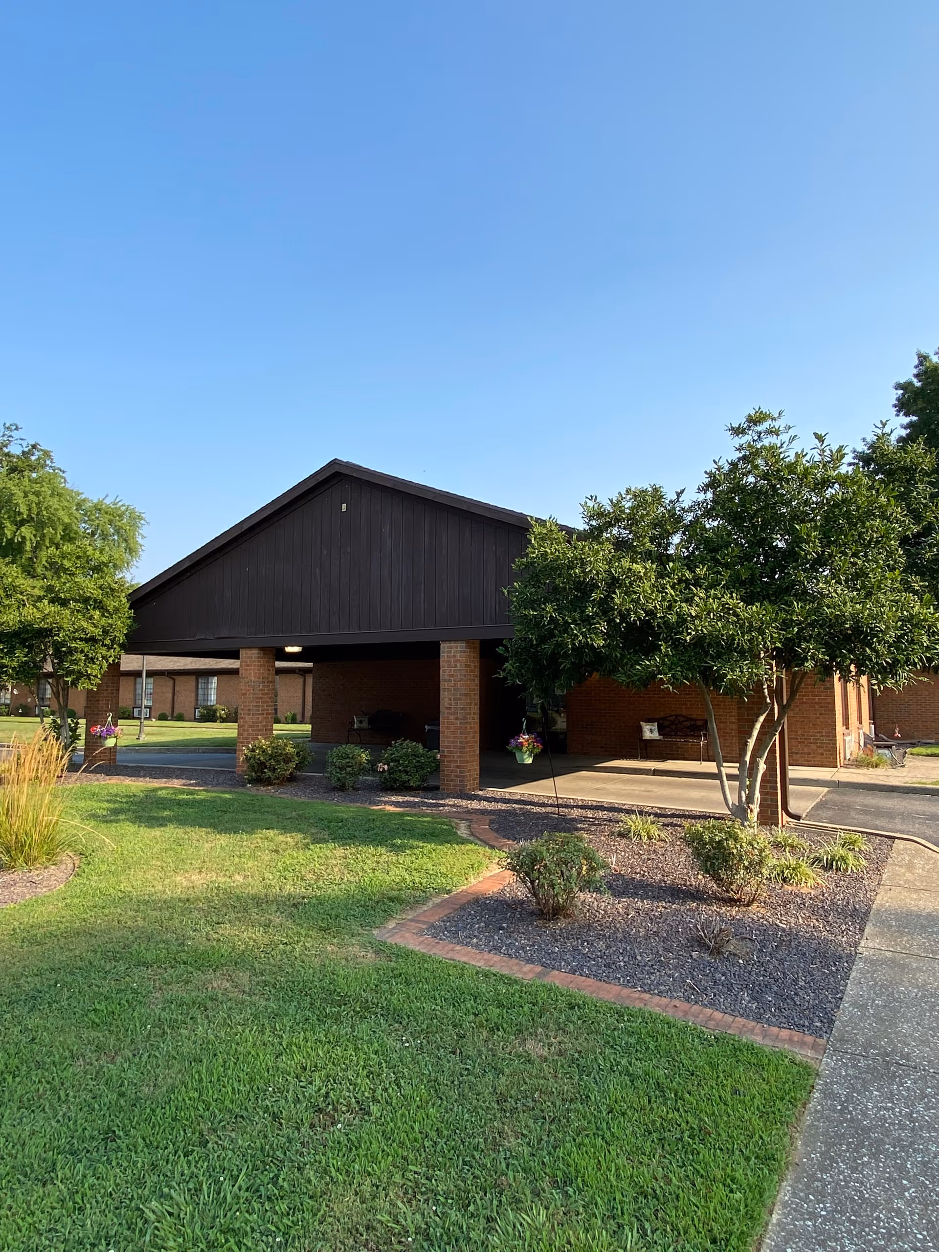 Covered brick entrance canopy of a single-story care facility with a manicured lawn and trees under a clear blue sky.