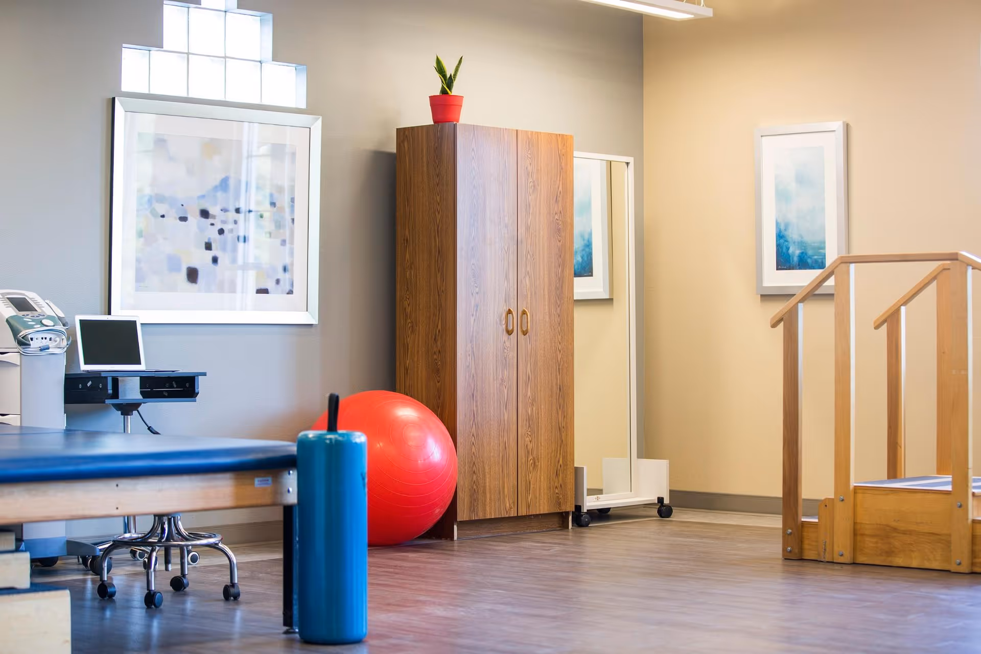 A bright therapy/exercise room with a treatment table, red exercise ball, wooden cabinet, mirror, and a small set of wooden steps.