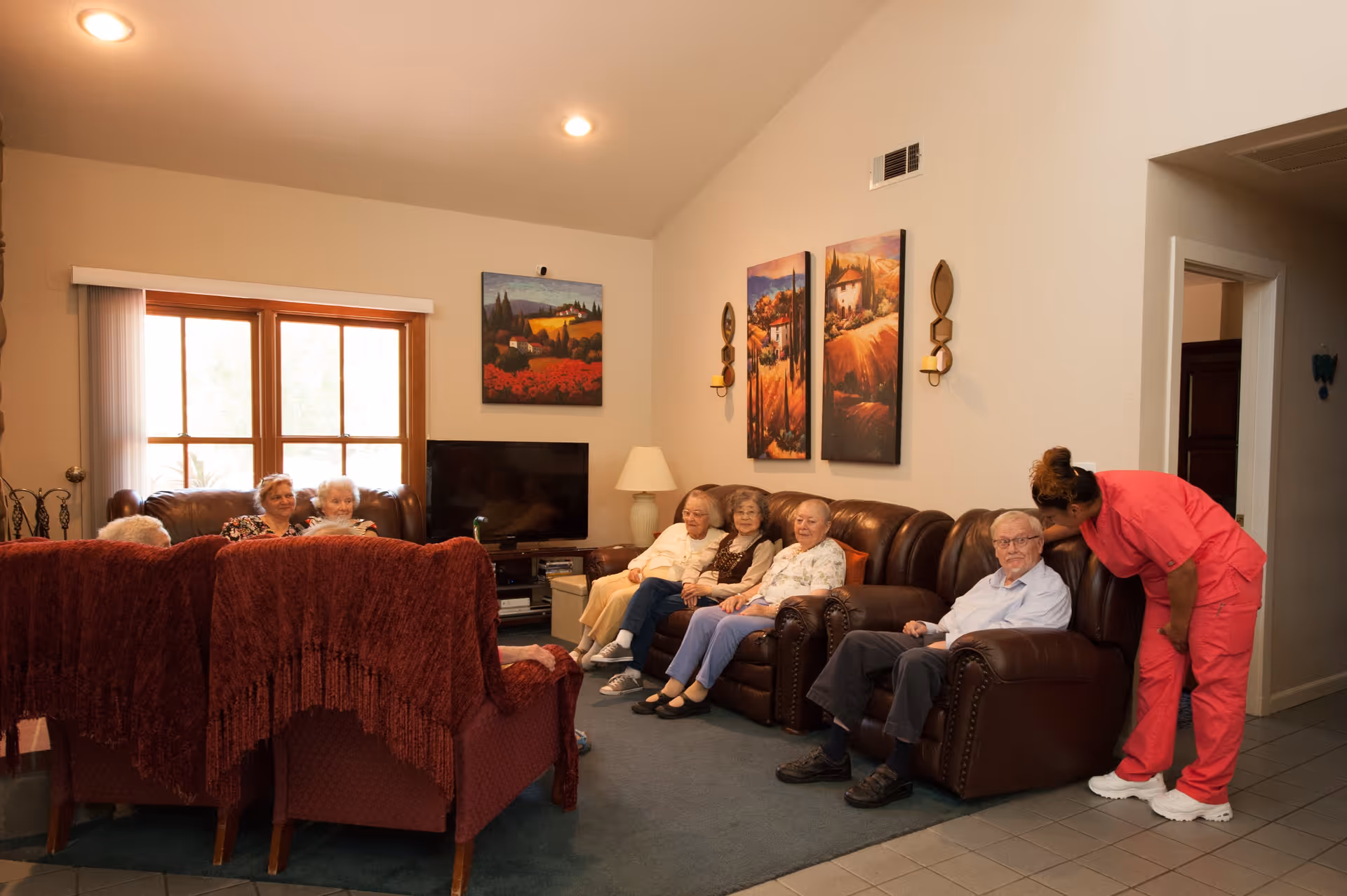 A group of elderly people sitting on leather sofas and chairs in a cozy living room with a caregiver in pink scrubs interacting with one of the men. The room has a large window with vertical blinds, a TV, and colorful landscape paintings on the wall.