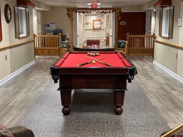 Indoor recreational area with a red felt pool table centered in the room. The pool table has two crossed cues and a rack of billiard balls arranged at one end. The room has light-colored walls, wood flooring, and windows with brown valances on both sides. In the background, there is a seating area with a red cushioned chair and decorative plants.