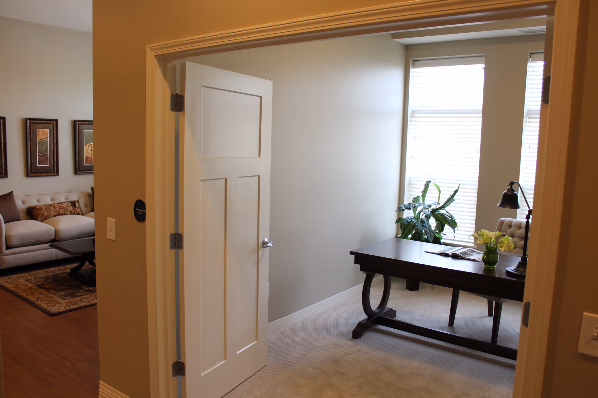 View through an open door into a small office with a dark wooden desk, a chair, a desk lamp, a vase with flowers, and a plant near two windows with blinds. To the left, part of a living room with a beige sofa, coffee table, and framed artwork on the wall is visible.