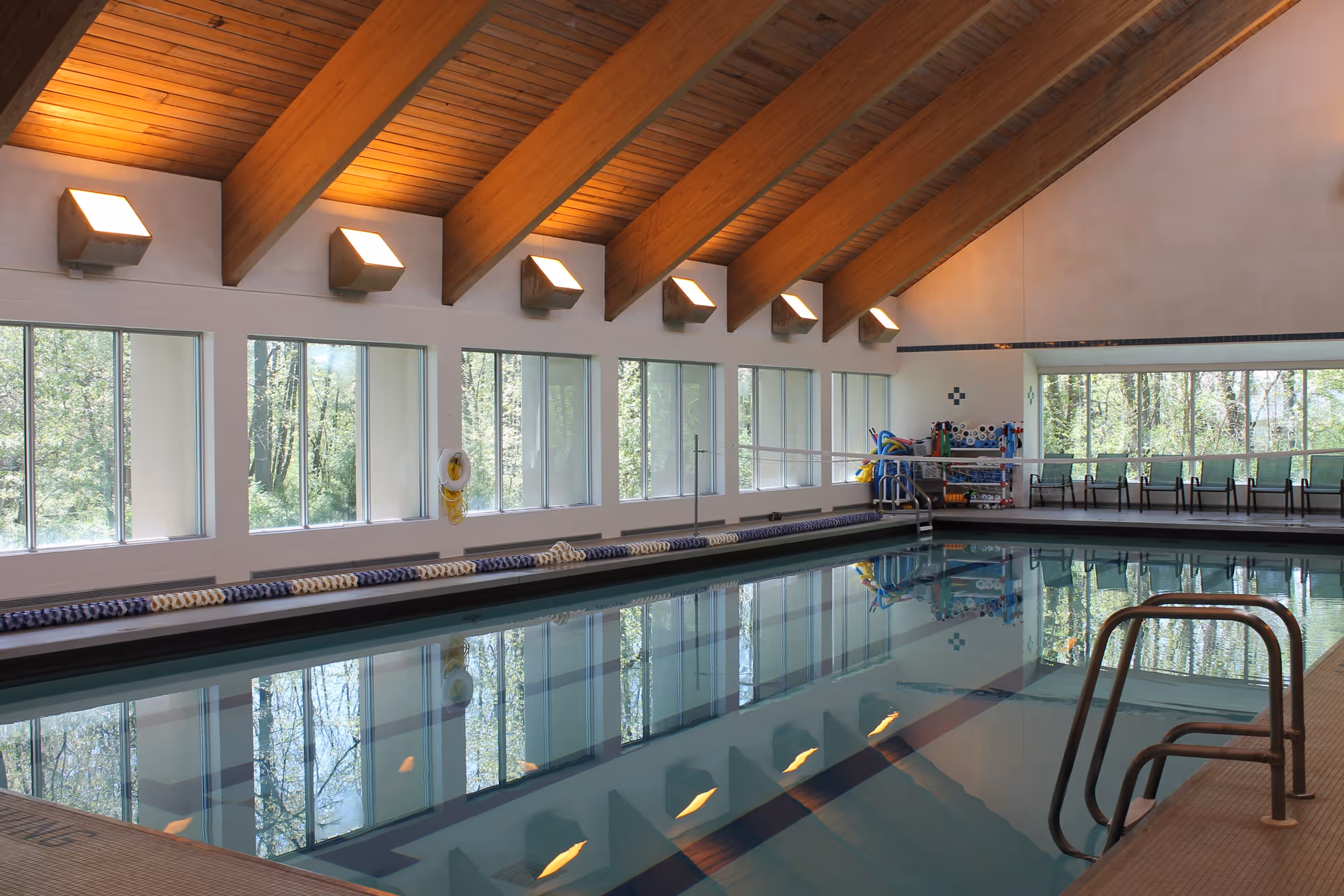 Indoor swimming pool with clear water, wooden beam ceiling, large windows letting in natural light, poolside chairs, and pool equipment stored at the far end.
