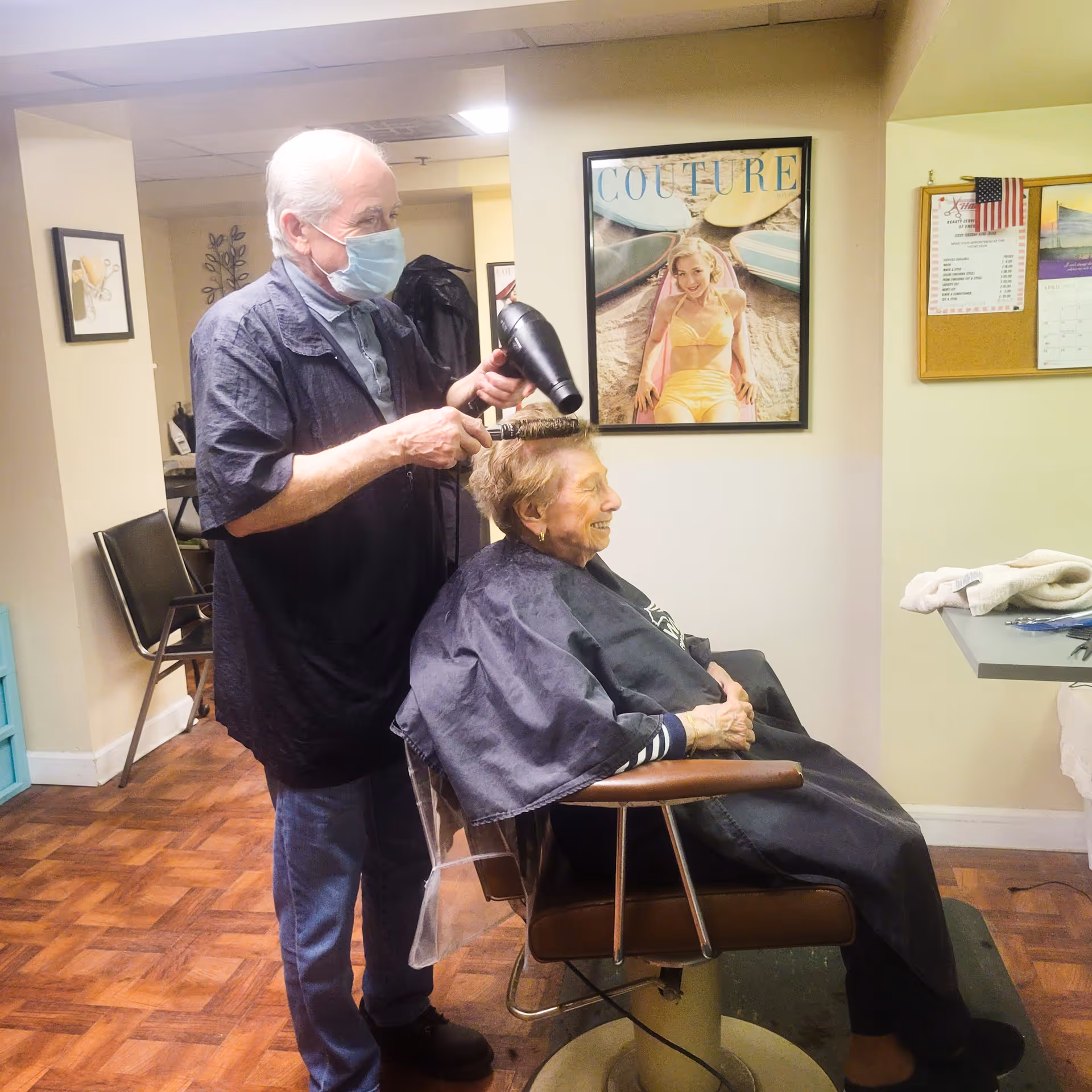 An elderly woman sits in a salon chair wearing a black cape while a man wearing a face mask blow-dries her hair in a senior living community salon. The room has wooden flooring, framed pictures on the walls, and a bulletin board with an American flag.