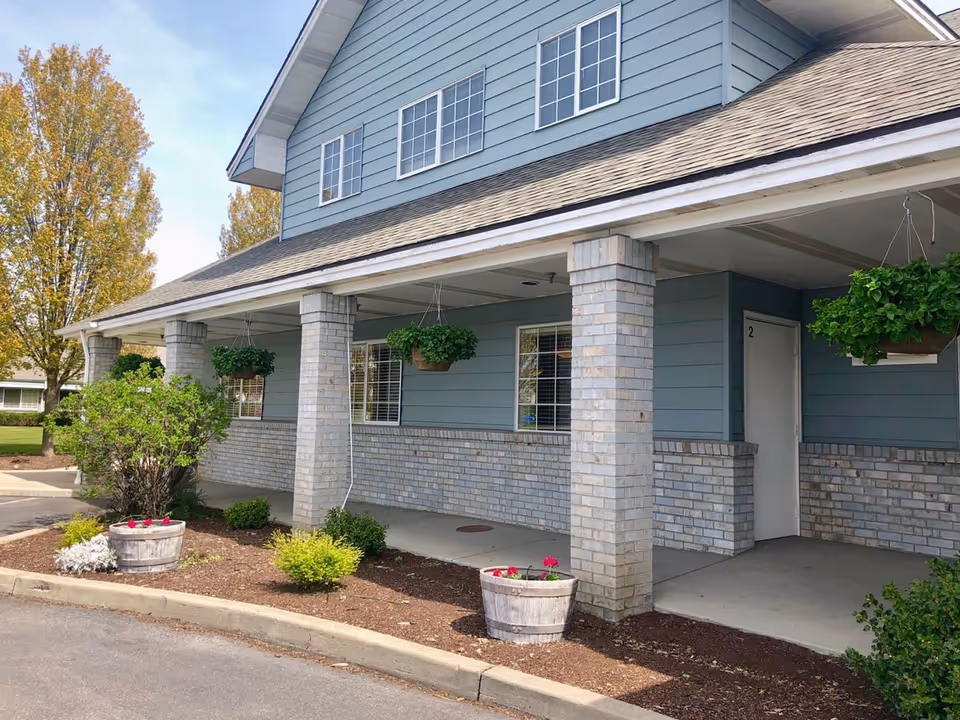 Exterior view of a two-story assisted living facility building with light blue siding and brick columns supporting a covered porch. There are hanging flower baskets and potted plants along the walkway, with trees and a clear sky in the background.