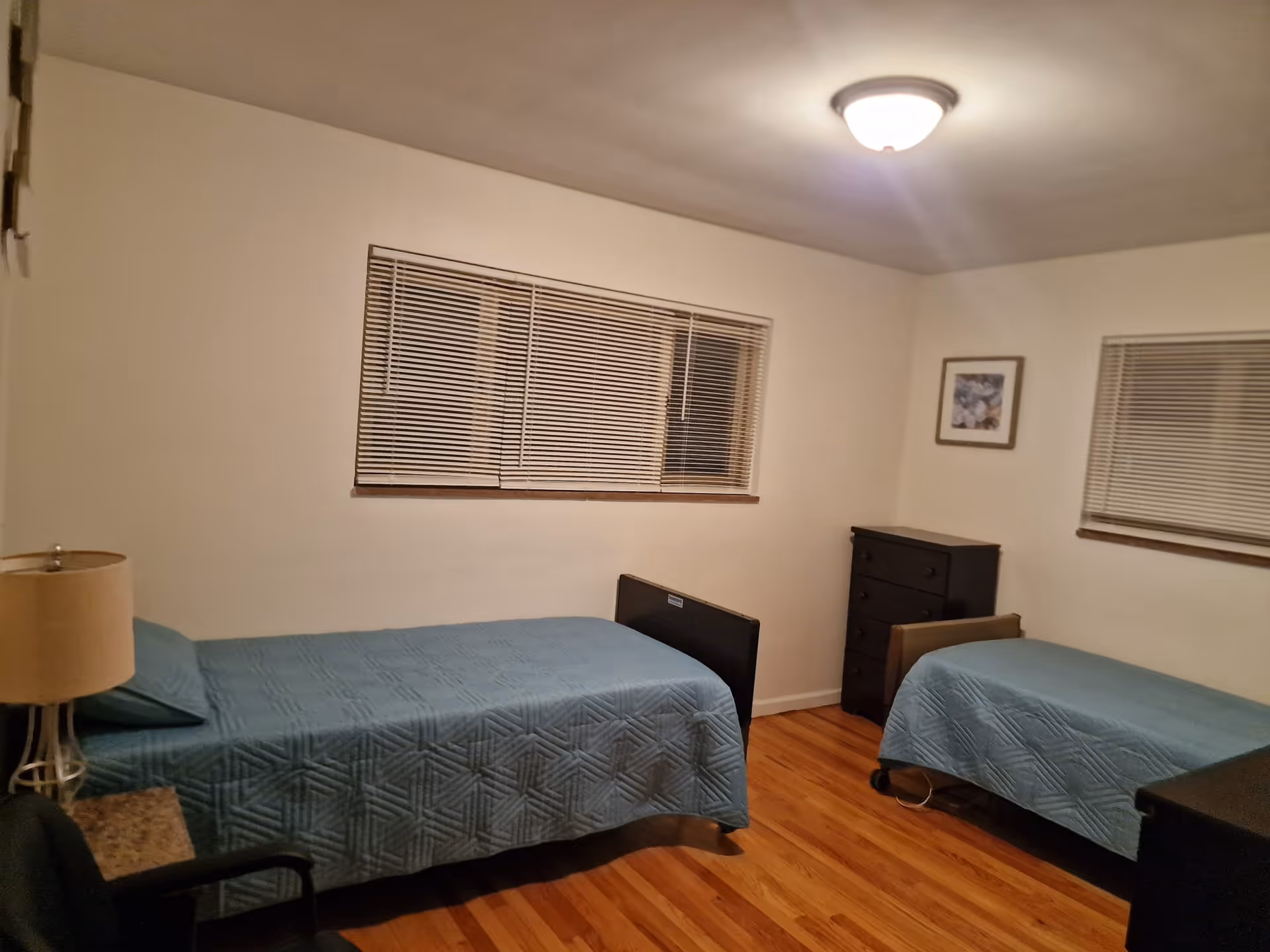 A simple bedroom with two single beds covered with blue quilts, a wooden floor, two windows with blinds, a small dresser, a lamp on a side table, and a framed picture on the wall.