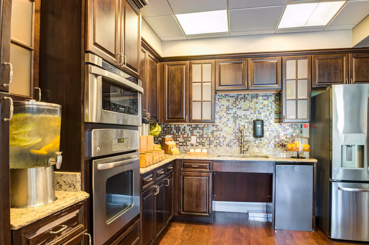 A modern kitchen with dark wooden cabinets, stainless steel double oven, refrigerator, and a mosaic tile backsplash. The countertop has a glass dispenser with lemon water, a fruit basket with bananas, and a cake stand with muffins. The floor is wooden and the ceiling has recessed lighting.