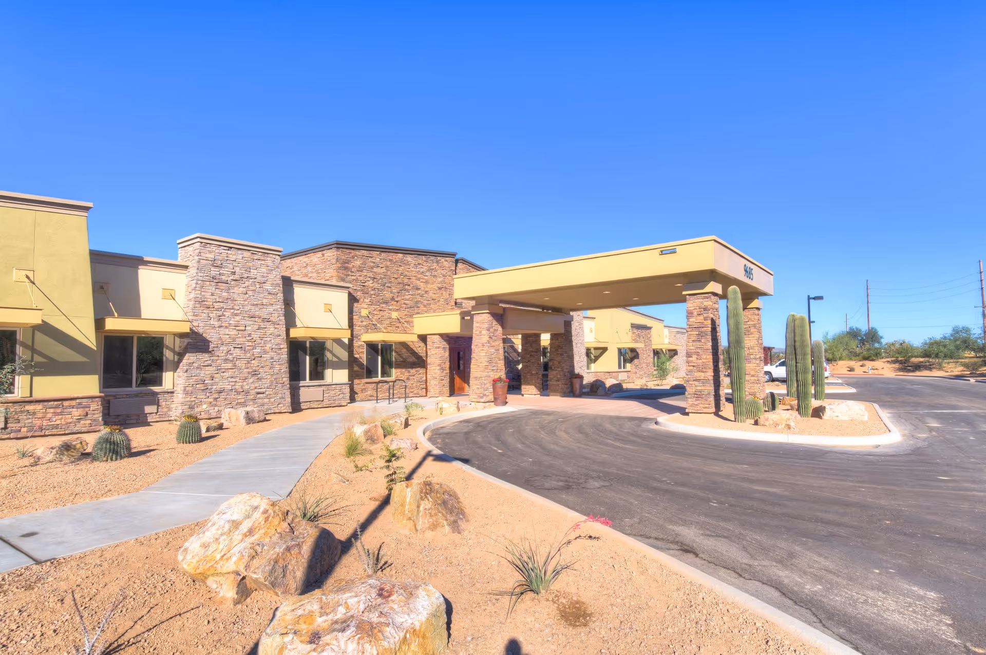 Exterior view of Catalina Springs Memory Care facility showing a single-story building with stone and stucco facade, a covered entrance supported by stone pillars, desert landscaping with rocks and cacti, and a clear blue sky.