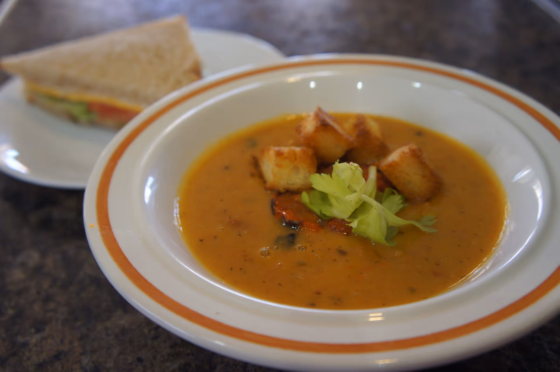 Bowl of creamy soup topped with croutons and celery with a half sandwich on a plate in the background.