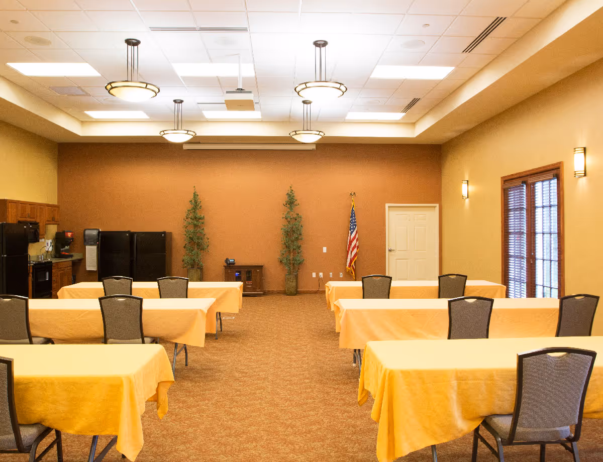 Large multipurpose dining/meeting room with rows of tables covered in yellow tablecloths, chairs, a kitchenette along the back wall, and an American flag.