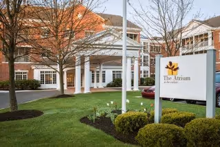 Exterior view of The Atrium At the Cedars senior living facility showing a brick building with white columns at the entrance, landscaped green lawn with bushes and trees, and a sign with the facility's name and logo.