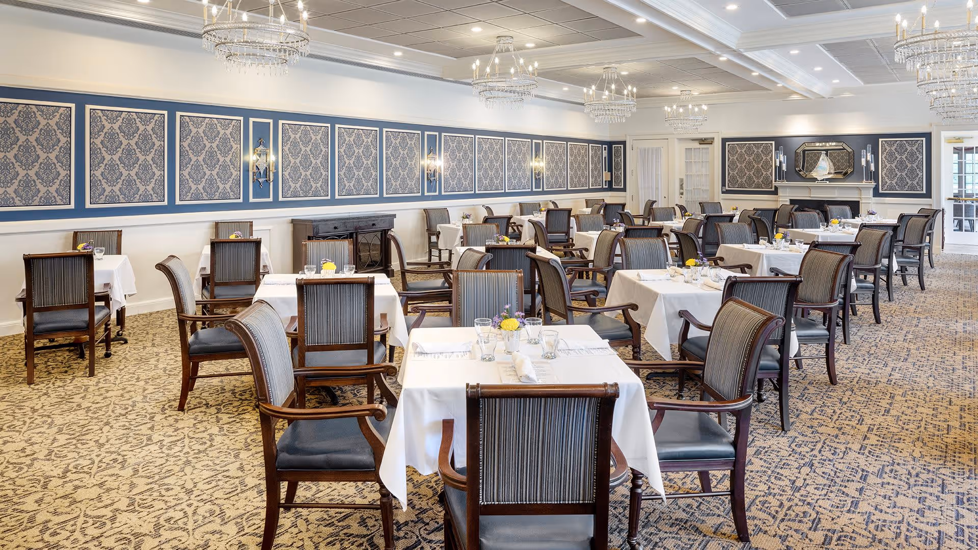 A spacious, elegant dining room with multiple tables covered in white tablecloths, each set with glassware, napkins, and small floral centerpieces. The room features patterned carpet, blue and beige damask wallpaper panels, wooden chairs with striped upholstery, chandeliers hanging from a coffered ceiling, and a fireplace with a decorative mirror above it.