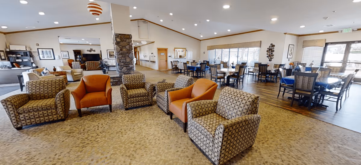 Spacious communal lounge and dining area with patterned armchairs grouped on carpet, dining tables on wood flooring, a stone column, and large windows.