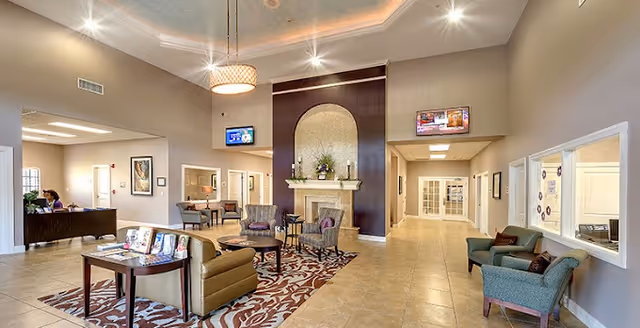 Spacious and well-lit senior living facility common area with comfortable seating including armchairs and sofas arranged around a patterned rug. A decorative fireplace with a floral arrangement is centered on a dark accent wall. Two wall-mounted televisions are visible above the fireplace and hallway entrance. Reception desk with staff is on the left side, and hallways lead to other rooms. The ceiling features recessed lighting and a hanging light fixture.