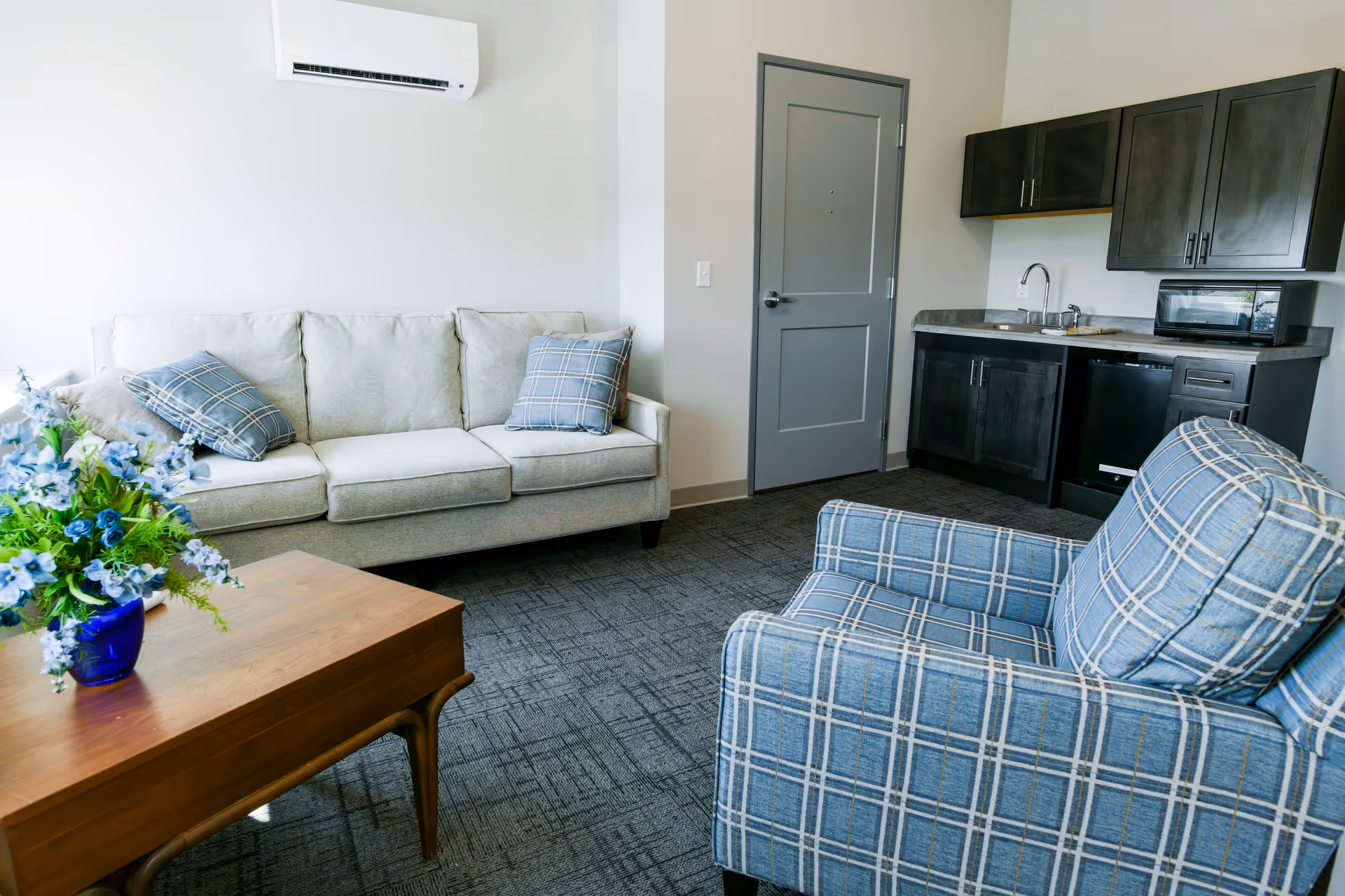 A cozy living area in an assisted living facility featuring a light gray sofa with two blue plaid pillows, a matching blue plaid armchair, a wooden coffee table with a blue vase holding blue and white flowers, a gray door, and a kitchenette with dark cabinets, a sink, a microwave, and a mini fridge.