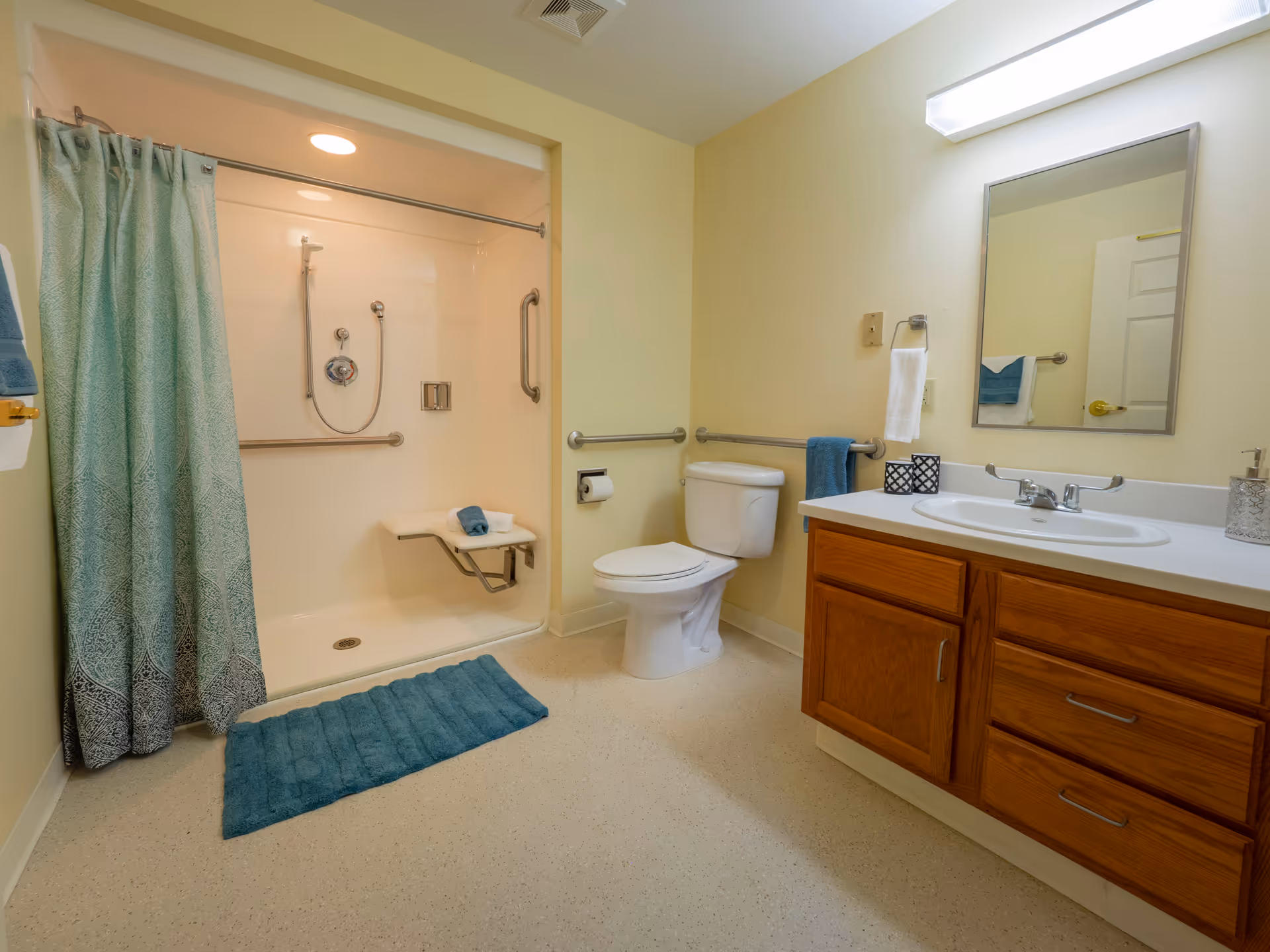 A clean and accessible bathroom featuring a walk-in shower with a fold-down seat and grab bars, a toilet with grab bars on both sides, a wooden vanity with a sink, a mirror above the sink, and a blue bath mat on the floor. The walls are painted light yellow and the shower has a blue patterned curtain.