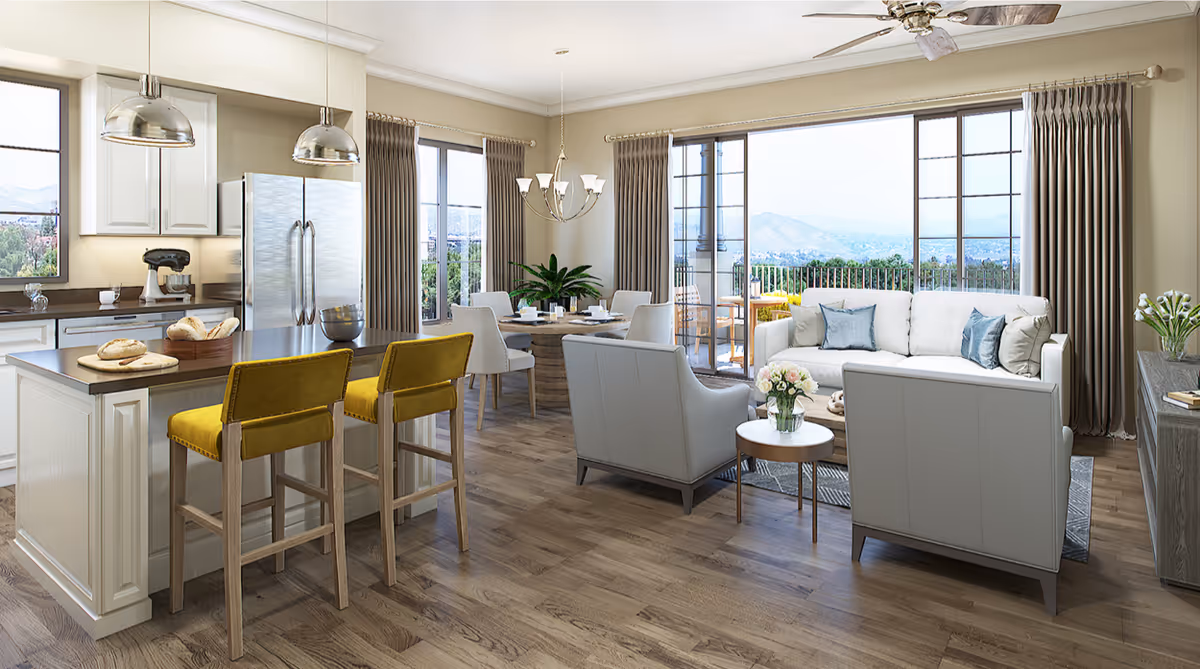 A bright and spacious open-concept living area featuring a modern kitchen with stainless steel refrigerator, island with two yellow bar stools, a dining table with six chairs, and a cozy living room with white sofa and two armchairs. Large windows and glass doors offer a scenic view of mountains and greenery outside.