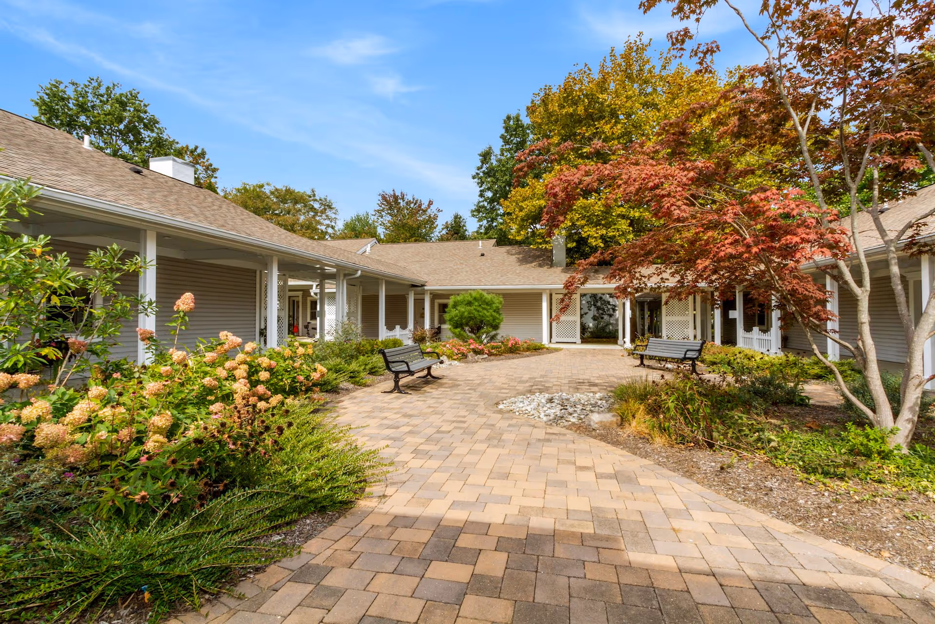 Paved courtyard with benches, landscaped plantings, and a covered walkway between single-story buildings under a blue sky.