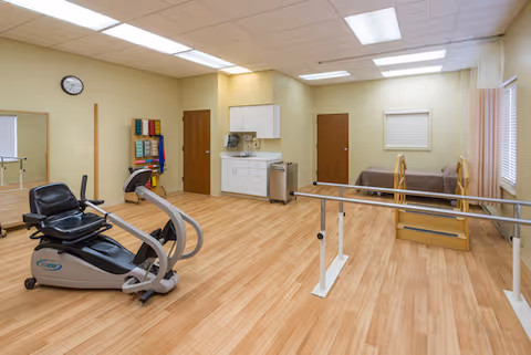 Therapy/exercise room with a recumbent exercise bike, parallel bars, treatment table, cabinets, and wall-mounted exercise equipment.