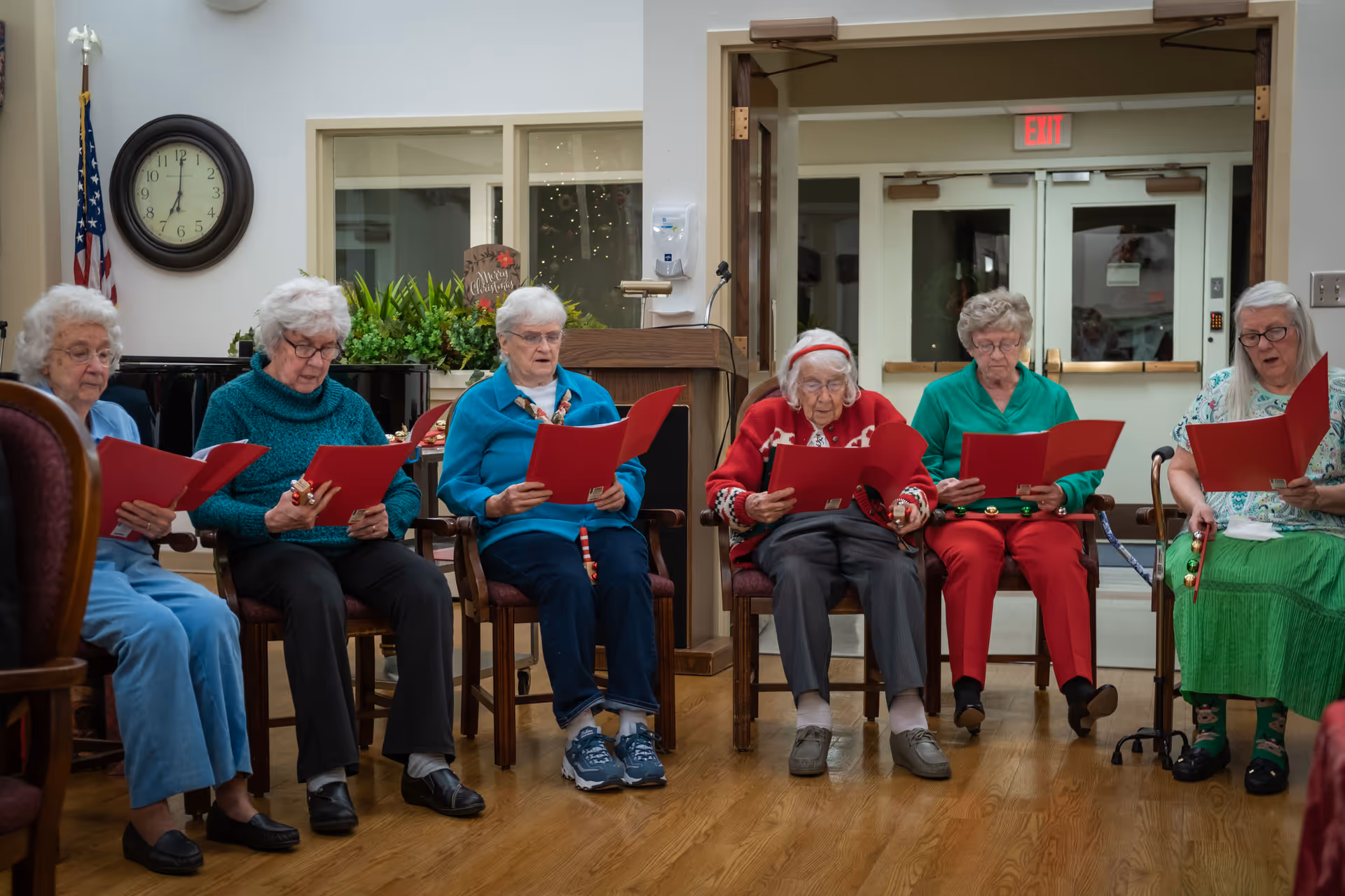 A group of elderly women seated in a communal room holding red songbooks and singing.