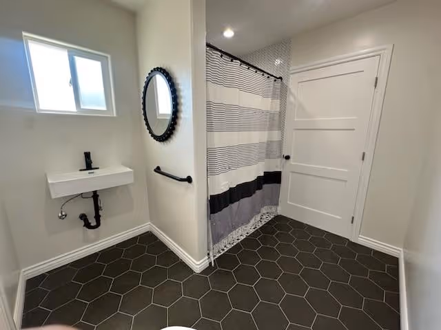 A modern bathroom with dark hexagonal floor tiles, a small white wall-mounted sink with black faucet and exposed plumbing, a round black-framed mirror above the sink, a black towel bar on the wall, a window with frosted glass, a shower area with a striped shower curtain, and a white door.