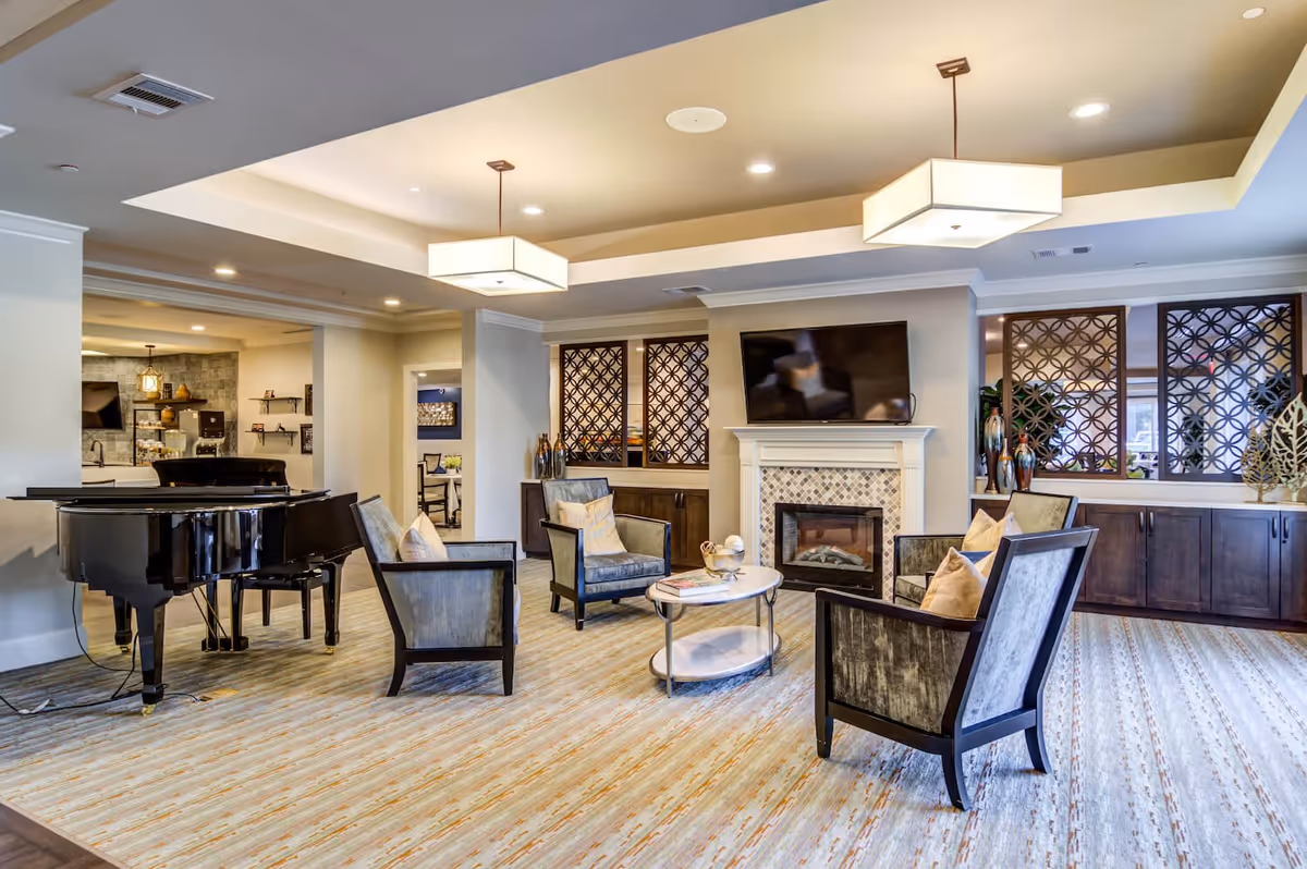 A spacious and well-lit living room area in Arbor Terrace Burnt Hickory featuring four cushioned armchairs arranged around a round coffee table. A fireplace with a mounted flat-screen TV above it is centered on the far wall. To the left, there is a black grand piano with a piano bench. The room has a light-colored carpet with a subtle pattern, recessed lighting, and two modern rectangular ceiling light fixtures. Decorative wooden lattice panels and cabinets line the back wall.
