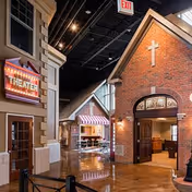 Interior view of a senior living facility designed to resemble a small town street with building facades, including a brick building with a cross above the door and a theater entrance with a marquee sign. The area has polished floors, overhead lighting, and a cozy, welcoming atmosphere.