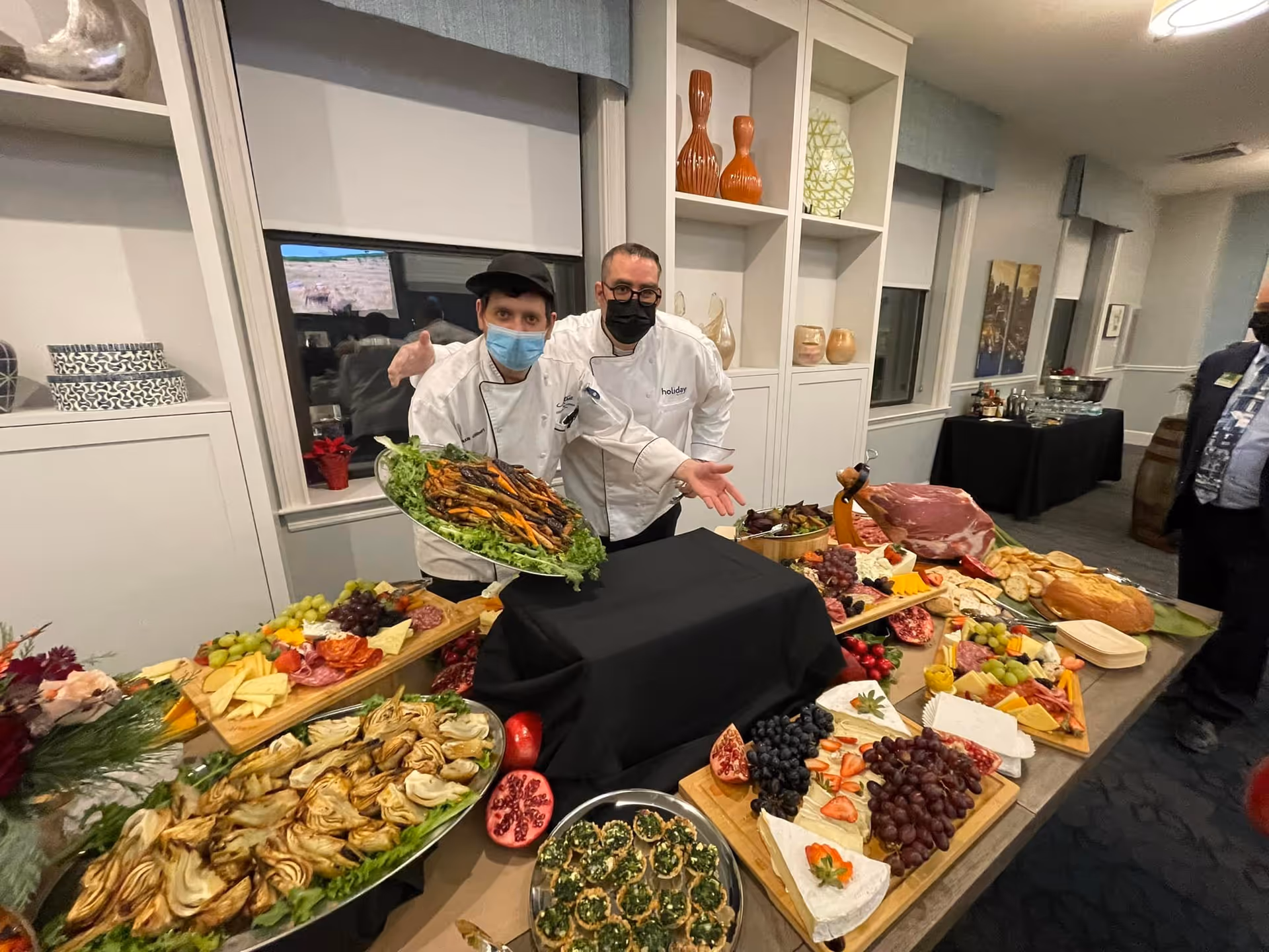 Two chefs wearing white uniforms and face masks stand behind a table filled with an assortment of food including grilled vegetables, cheeses, grapes, sliced meats, bread, and various appetizers in a well-lit room with shelves and windows.