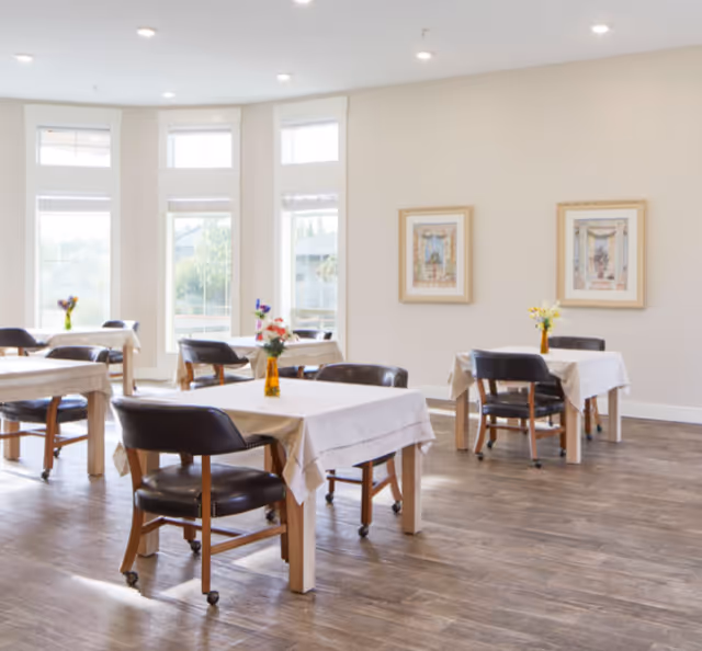 Bright dining area with multiple small tables covered in white tablecloths, leather chairs, vases of flowers, and large windows.