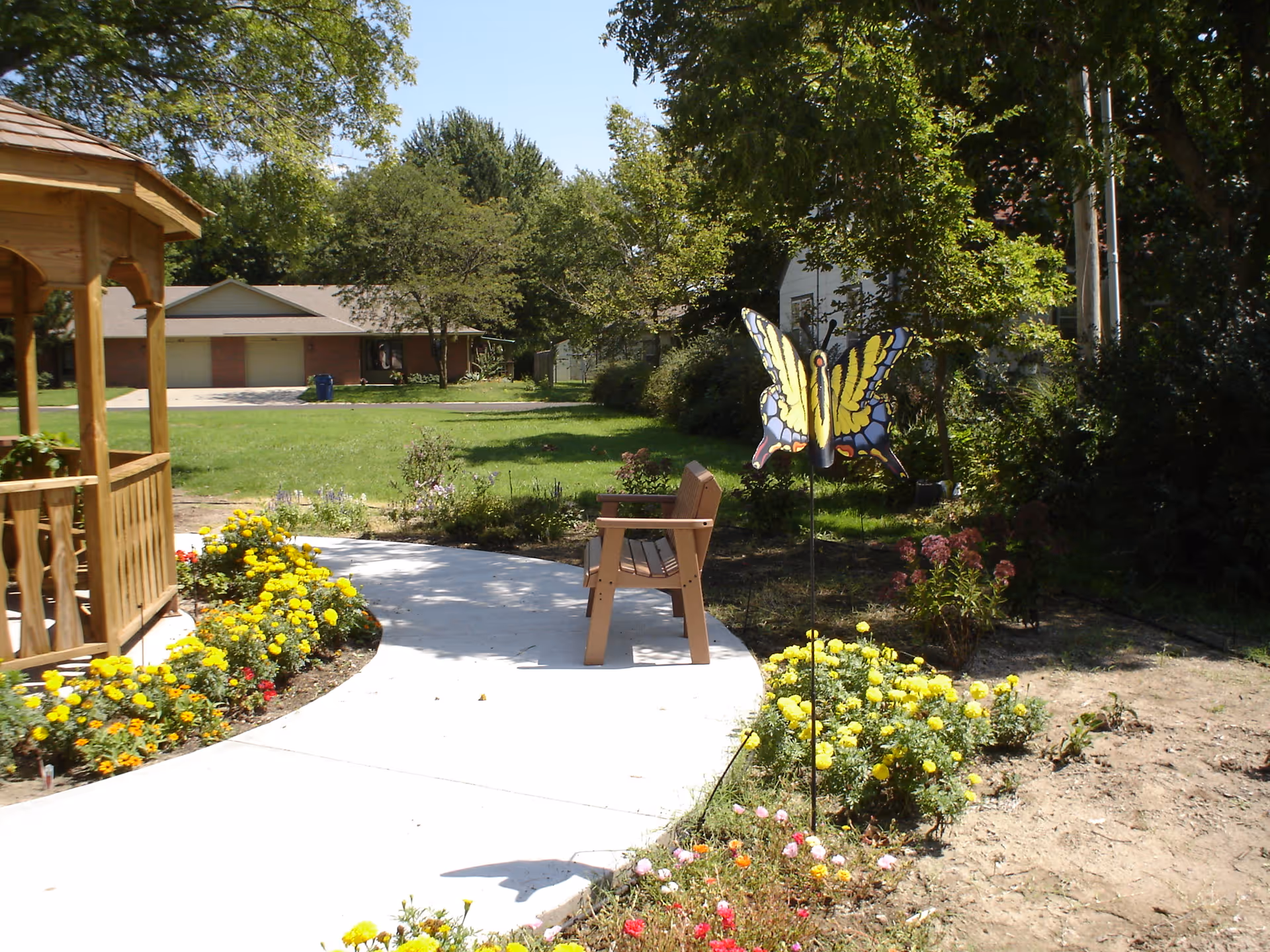 A sunny outdoor garden area with a concrete pathway, a wooden bench, colorful flower beds, a wooden gazebo on the left, and a decorative butterfly garden stake. Trees and houses are visible in the background.