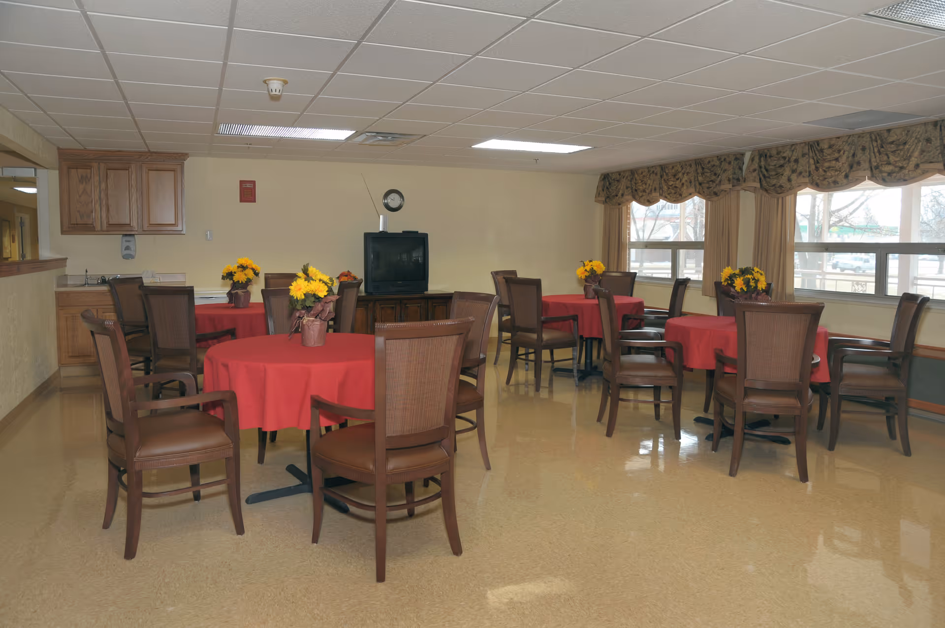 Communal dining room with round tables draped in red tablecloths, wooden chairs, floral centerpieces, and a TV against the far wall.