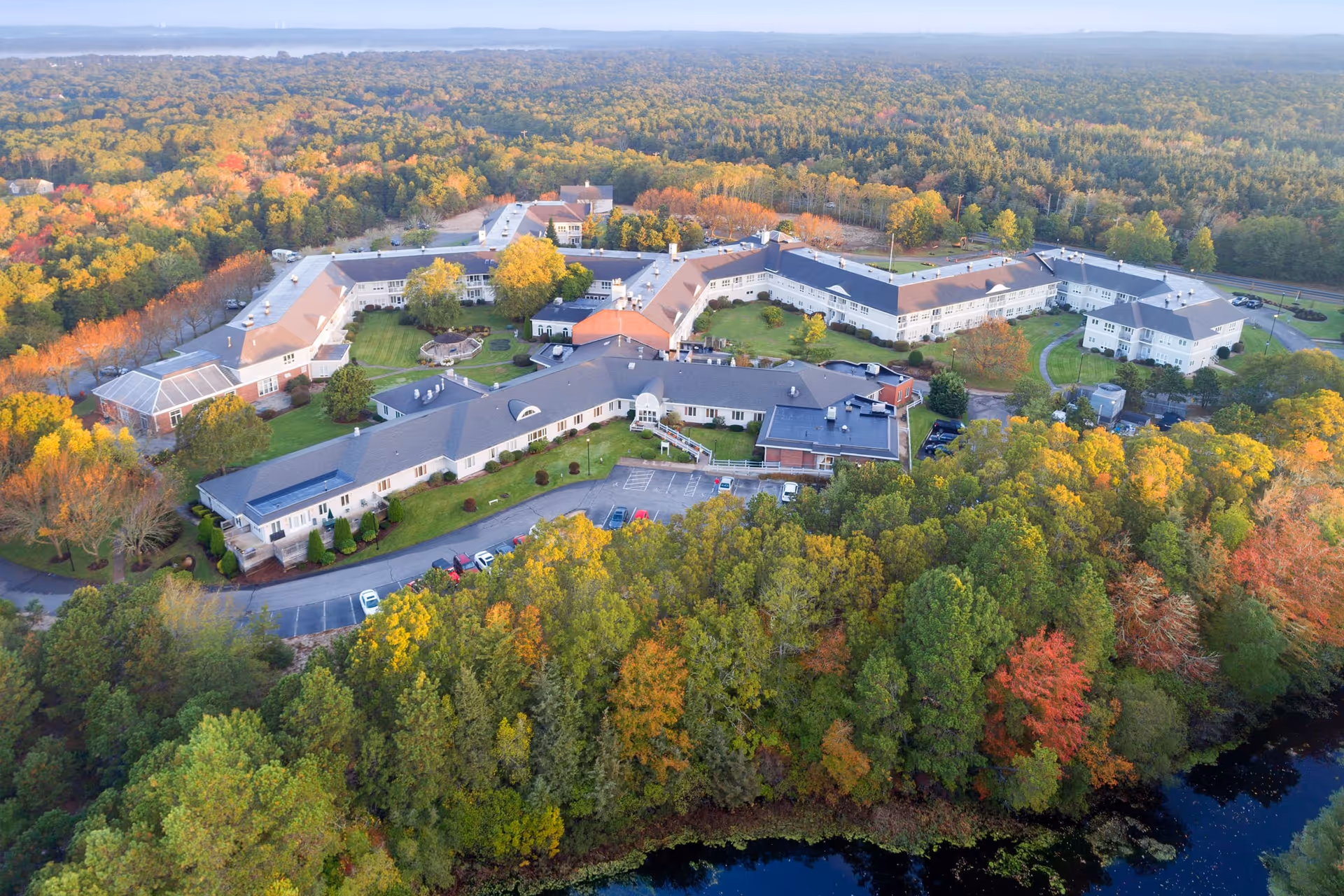 Aerial view of Maplewood at Mayflower Place, a large senior living facility surrounded by dense trees with autumn foliage. The facility consists of multiple connected buildings with parking areas and green lawns, set in a forested landscape with a body of water visible at the bottom right.
