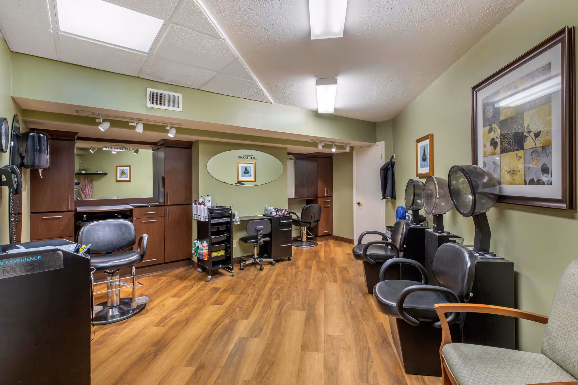 Interior view of a hair salon area in a senior living facility with green walls and wood flooring. The room features salon chairs, hair drying stations, a large mirror, cabinets, and framed artwork on the walls. There is also a small rolling cart with hair products and tools.