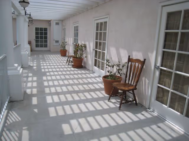 Sunlit covered exterior corridor with potted plants and wooden chairs beside French doors casting grid-like shadows on the floor.