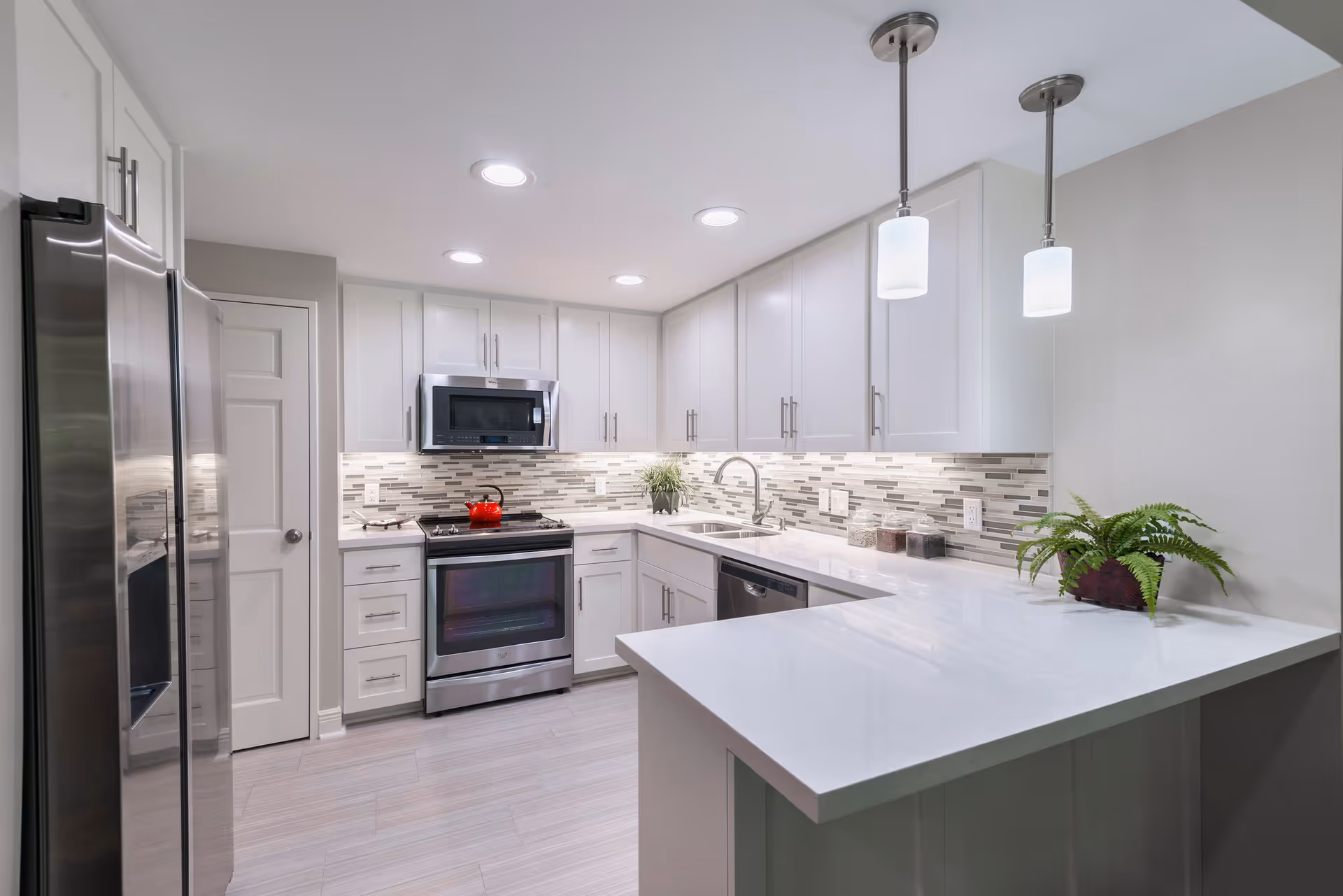 Modern kitchen with white cabinets, stainless steel refrigerator, oven, microwave, and dishwasher. The kitchen features a white countertop with a plant and jars, a tiled backsplash, recessed ceiling lights, and two pendant lights hanging above the counter.