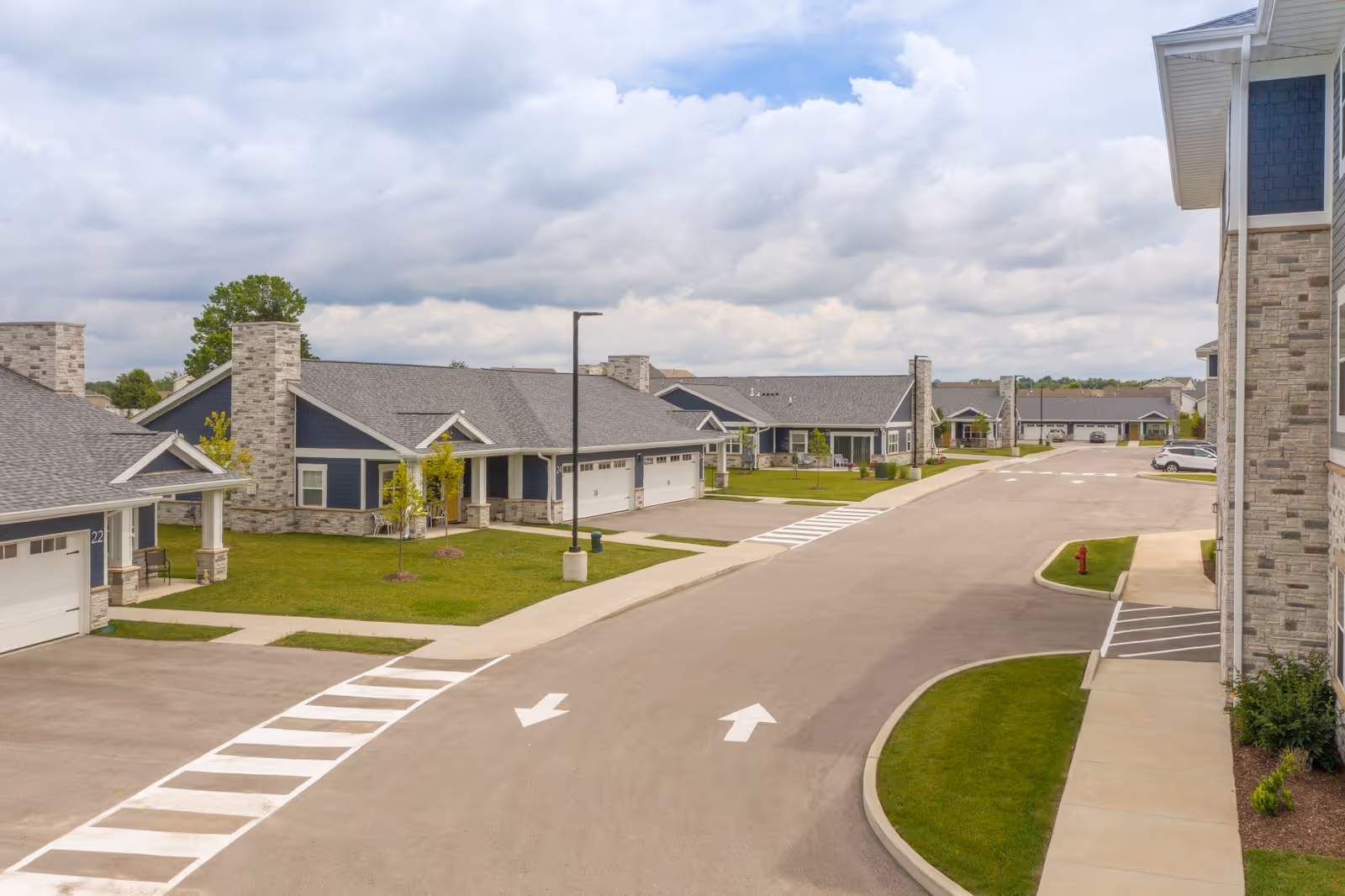 View of a residential street within a senior living community featuring single-story homes with stone and blue siding, garages, small lawns, and a cloudy sky overhead.