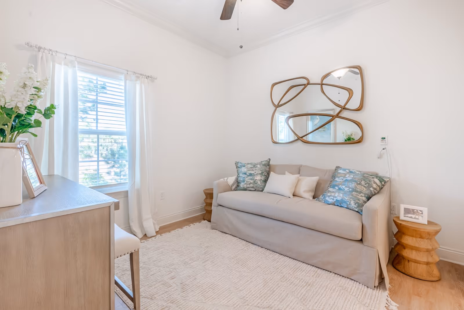 A bright and cozy living room with a beige sofa adorned with patterned and plain pillows, a unique abstract wall mirror above the sofa, a wooden side table with a framed photo, a light-colored wooden desk with a chair, and a window with white curtains letting in natural light.
